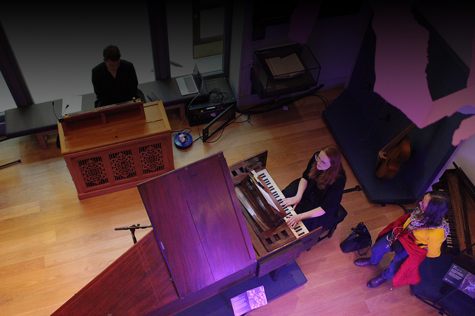 Students performing on a harpsichord, surrounded by antique instruments and furniture in the museum.